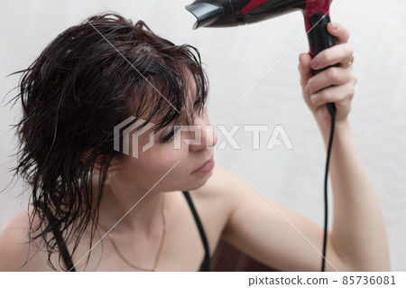 A woman after a shower dries her hair by the mirror with a hairdryer A woman after a shower dries her hair by the mirror with a hairdryer 85736081