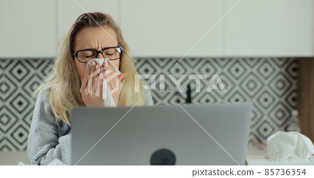 Close up of sick woman blows her nose into a paper napkin while working remotely at home. Concept of healthcare and illness. Close up of sick woman blows her nose into a paper napkin while working remotely at home. Concept of healthcare and illness. 85736354