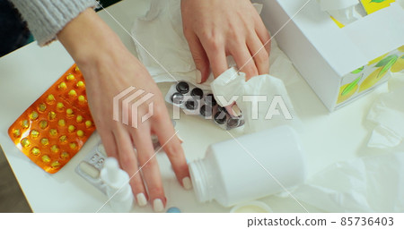 Close up of hand holding tablets with capsules and pills against cold and flu. Table with medicaments, bottle with medicine, thermometer, tissues for sick person. 85736403