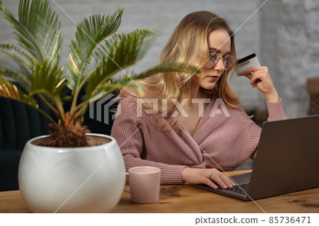 Girl in pink cardigan, glasses. Working on laptop and showing white plastic card. Sitting at wooden table with pink cup, palm in pot on it. Close up Girl in pink cardigan, glasses. Working on laptop and showing white plastic card. Sitting at wooden table with pink cup, palm in pot on it. Close up 85736471