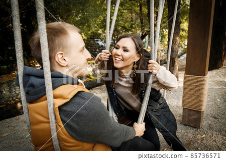 Cheerful young mixed race couple looking at each other while sitting on a swing outdoor Cheerful young mixed race couple looking at each other while sitting on a swing outdoor 85736571