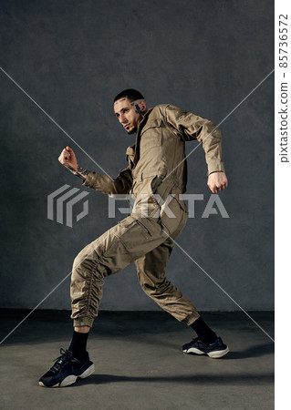Good-looking man with tattooed body, earrings, beard. Dressed in khaki overalls and black sneakers. Dancing on gray background. Dancehall, hip-hop Good-looking man with tattooed body, earrings, beard. Dressed in khaki overalls and black sneakers. Dancing on gray background. Dancehall, hip-hop 85736572