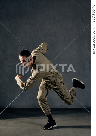 Stately fellow with tattooed body and face, earrings, beard. Dressed in khaki jumpsuit, black sneakers. Dancing on gray background. Dancehall, hip-hop Stately fellow with tattooed body and face, earrings, beard. Dressed in khaki jumpsuit, black sneakers. Dancing on gray background. Dancehall, hip-hop 85736576