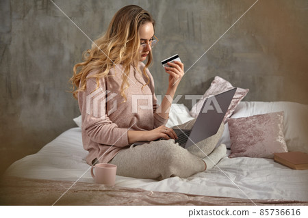 Blonde girl blogger in glasses, casual clothing. Holding plastic card and laptop on her legs, sitting sideways on bed with book and cup. Close up 85736616