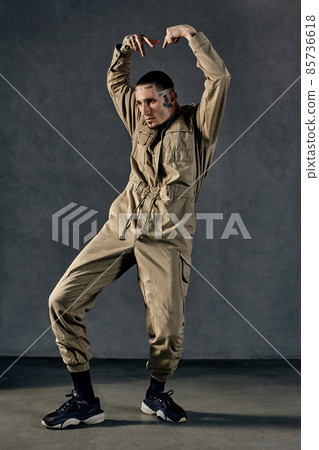 Active guy with tattooed body and face, earrings, beard. Dressed in khaki overalls and black sneakers. Dancing on gray background. Dancehall, hip-hop 85736618