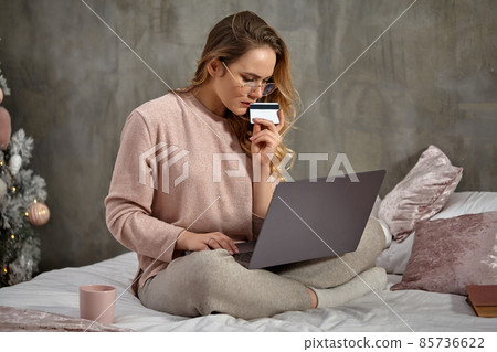 Blonde model blogger in glasses, casual clothing. Holding plastic card and working on her laptop, sitting on bed with book and cup. Close up Blonde model blogger in glasses, casual clothing. Holding plastic card and working on her laptop, sitting on bed with book and cup. Close up 85736622