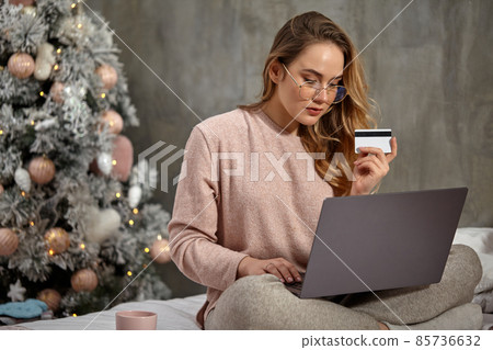 Blonde girl blogger in glasses, casual clothing. She is holding plastic card and working on her laptop, sitting on bed with cup. Close up, copy space Blonde girl blogger in glasses, casual clothing. She is holding plastic card and working on her laptop, sitting on bed with cup. Close up, copy space 85736632