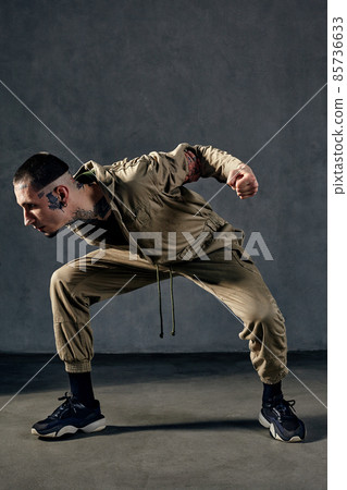 Unusual man with tattooed body and face, earrings, beard. Dressed in khaki overalls and black sneakers. Dancing on gray background. Dancehall, hip-hop Unusual man with tattooed body and face, earrings, beard. Dressed in khaki overalls and black sneakers. Dancing on gray background. Dancehall, hip-hop 85736633