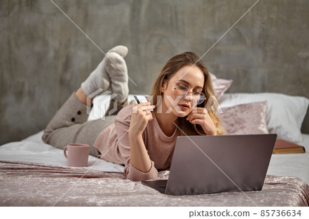 Blonde lady blogger in glasses, casual clothing. Holding white plastic card, lying on bed with laptop, book and cup. Close-up, copy space Blonde lady blogger in glasses, casual clothing. Holding white plastic card, lying on bed with laptop, book and cup. Close-up, copy space 85736634
