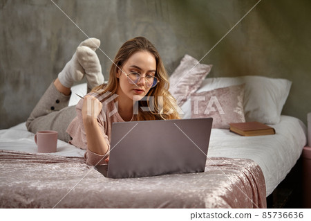 Blonde model blogger in glasses, casual clothing. Holding white plastic card, lying on bed with laptop, book and cup. Work or education. Close-up Blonde model blogger in glasses, casual clothing. Holding white plastic card, lying on bed with laptop, book and cup. Work or education. Close-up 85736636