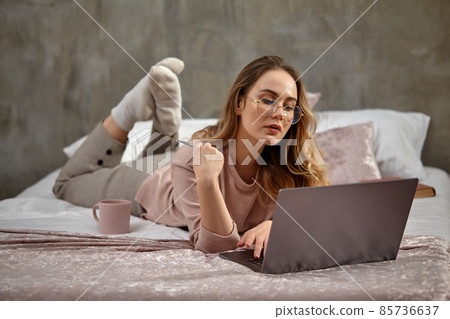 Blonde woman blogger in glasses, casual clothing. Holding plastic card, lying cross-legged on bed with laptop, book and cup. Work, education. Close-up Blonde woman blogger in glasses, casual clothing. Holding plastic card, lying cross-legged on bed with laptop, book and cup. Work, education. Close-up 85736637