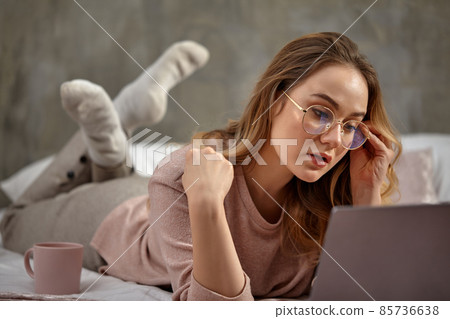 Blonde female blogger in glasses, casual clothing. Lying cross-legged on bed with cup, looking at her laptop. Interior with gray wall. Close up 85736638