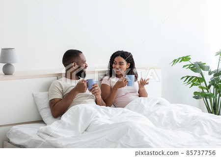 Cheerful young black husband and wife relaxing in bed with cups of coffee and talking, enjoy morning together Cheerful young black husband and wife relaxing in bed with cups of coffee and talking, enjoy morning together 85737756