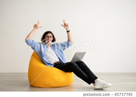 Happy freelance guy sitting in beanbag chair with brand new laptop, pointing up at copy space over light wall, panorama Happy freelance guy sitting in beanbag chair with brand new laptop, pointing up at copy space over light wall, panorama 85737795
