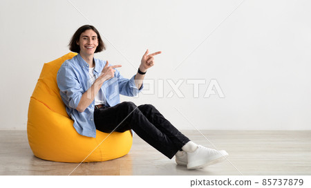 Positive caucasian guy pointing fingers aside at free space, sitting in beanbag chair over light wall, panorama 85737879