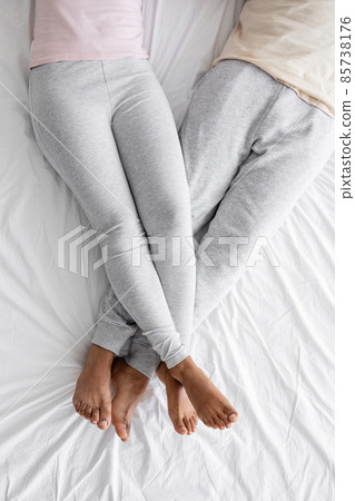 Cropped millennial african american woman in domestic clothes put her feet on man on white comfort bed Cropped millennial african american woman in domestic clothes put her feet on man on white comfort bed 85738176
