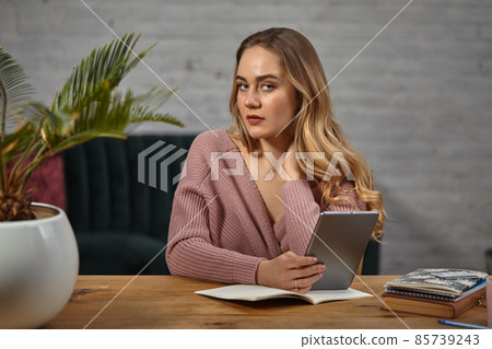 Female blogger in pink cardigan is holding a tablet. Sitting at wooden table with notebooks and palm tree in pot on it. Student, blogger. Close-up 85739243