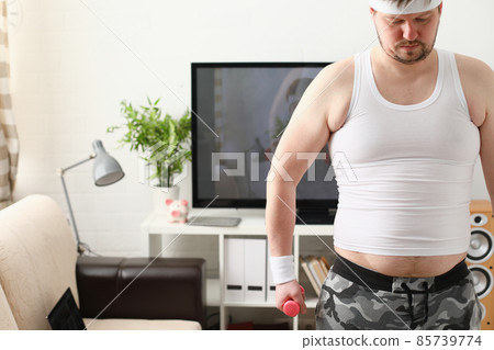 Overweight man is engaged in fitness at home holding dumbbell in hand 85739774