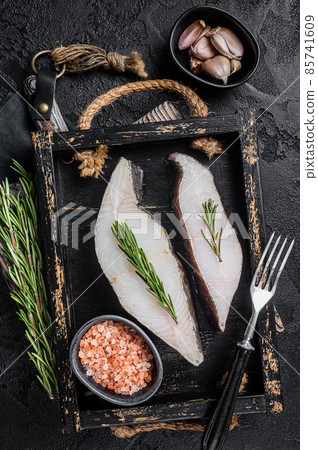 Raw halibut fish steak in wooden tray with herbs. Black background. Top view 85741609