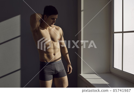 Young man posing in underwear. Male model with fit body in comfortable black cotton underpants standing near window. Studio shot with contrasting bright sunlight and shadows. Male underwear concept 85741987