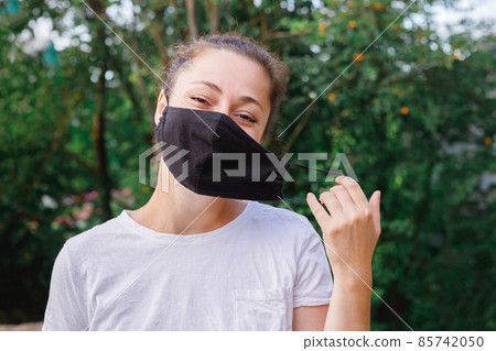 Happy positive girl takes off protective medical mask from face outdoors. Young woman removing mask smiling after vaccination. Coronavirus pandemic Covid 19 concept. Spring flowers pollen allergy 85742050