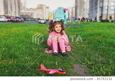 happy little girl in pink sportswear girl sitting on scooter on grass in city happy little girl in pink sportswear girl sitting on scooter on grass in city 85743132