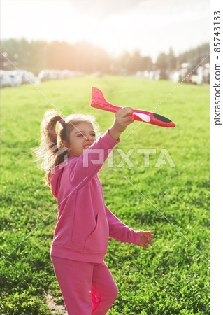 happy girl in pink sportswear with red toy plane running on green grass in sunny summer happy girl in pink sportswear with red toy plane running on green grass in sunny summer 85743133