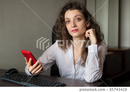 Tired young woman with curly hair and white shirt is working at the office and using her red smartphone, routine work, freelance, burnout syndrome 85743428