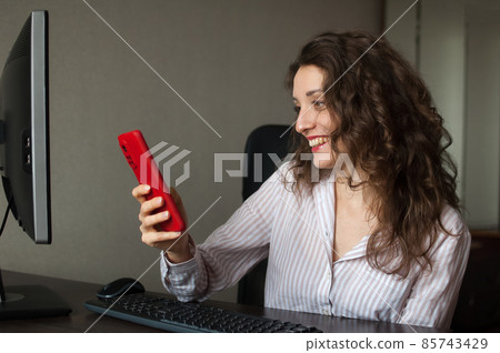 Tired young woman with curly hair and white shirt is working at the office and using her red smartphone, routine work, freelance, burnout syndrome Tired young woman with curly hair and white shirt is working at the office and using her red smartphone, routine work, freelance, burnout syndrome 85743429
