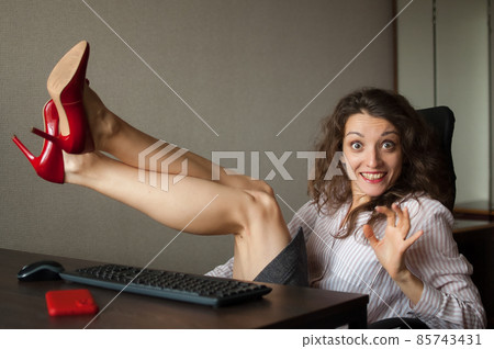 Portriat of brunette with legs on office table with laptop, hot woman in red shoes on high heels sitting at the desk. Concept of seductive secretary, female fashion, relax at work 85743431