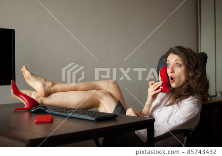 Crazy young woman is sitting at the desk with legs in red high heels on office table and pretending that she is talking by phone using one of her shoes 85743432
