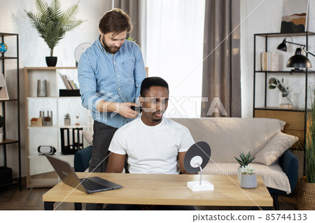 Bearded Caucasian man shaves a neck of African man with an electric razor sitting at the table 85744133