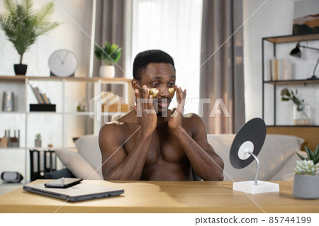 A man sits at a wooden table and apply patches under his eyes looking into the mirror. 85744199