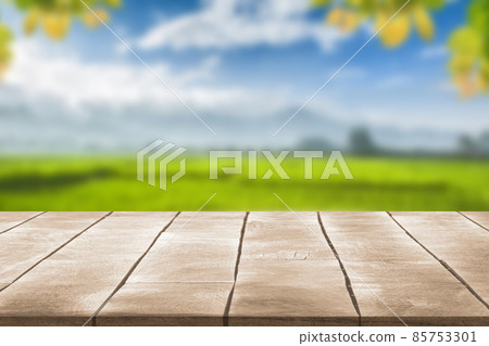 Wooden table top with blurry rice plantation landscape againt blue sky soft clouds with leaves frame Wooden table top with blurry rice plantation landscape againt blue sky soft clouds with leaves frame 85753301