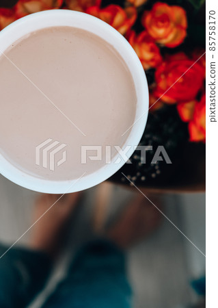Woman holding white cup of coffee, latte cappucino, in background of bouquet orange roses flowers. Good morning concept. Spring romance. Woman holding white cup of coffee, latte cappucino, in background of bouquet orange roses flowers. Good morning concept. Spring romance. 85758170