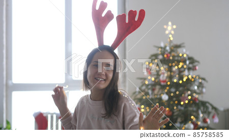 Young beautiful happy woman wearing reindeer antlers headband and galand dancing next to the Christmas tree Young beautiful happy woman wearing reindeer antlers headband and galand dancing next to the Christmas tree 85758585