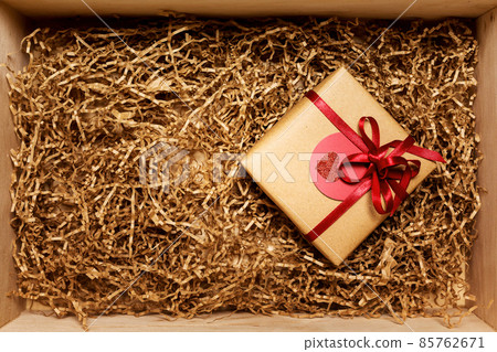 Wooden box with present wrapped in brown craft paper with red ribbon, filled with paper filler, top view Wooden box with present wrapped in brown craft paper with red ribbon, filled with paper filler, top view 85762671