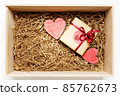 Wooden box with present wrapped in brown craft paper with red ribbon, filled with paper filler, top view 85762673