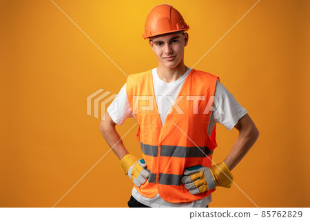 Confident smiling teen boy wearing orange hard hat against yellow background Confident smiling teen boy wearing orange hard hat against yellow background 85762829