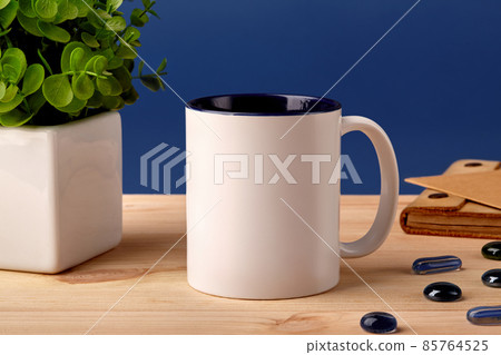 White ceramic mug on wooden desktop next to scattered glass stones, green plant in pot and brown notepad, blue background. Close up, copy space 85764525