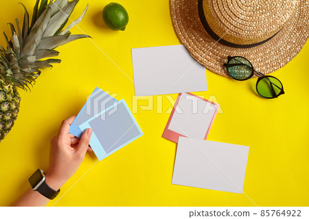 Hand of woman holding snapshot pictures. Straw hat, pineapple, lime, sunglasses and white sheets of paper on yellow background. Close up, copy space. Hand of woman holding snapshot pictures. Straw hat, pineapple, lime, sunglasses and white sheets of paper on yellow background. Close up, copy space. 85764922