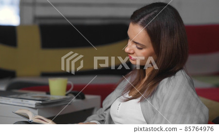 A young woman is sitting at home on an ottoman and reading a book. A woman is resting indoors on a comfortable ottoman in her free time. Have a nice free time on the weekend 85764976