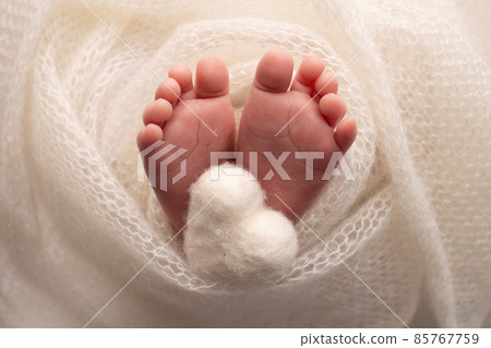 The tiny foot of a newborn baby. Soft feet of a new born in a white wool blanket. Close up of toes, heels and feet of a newborn. Knitted white heart in the legs of a baby. Macro photography. The tiny foot of a newborn baby. Soft feet of a new born in a white wool blanket. Close up of toes, heels and feet of a newborn. Knitted white heart in the legs of a baby. Macro photography. 85767759