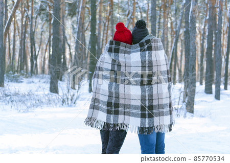 Guy and girl in knitted hats with buboes standing in winter in park are covered with warm blanket. Back view. Guy and girl in knitted hats with buboes standing in winter in park are covered with warm blanket. Back view. 85770534