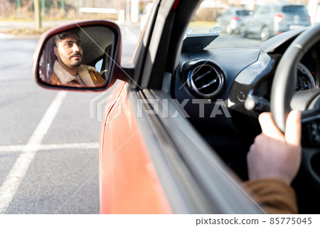 Man in jacket holding driving wheel riding car at daytime. Face in rear mirror Man in jacket holding driving wheel riding car at daytime. Face in rear mirror 85775045