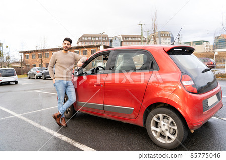 Young latin or arab eastern man standing near red small car on parking slot Young latin or arab eastern man standing near red small car on parking slot 85775046