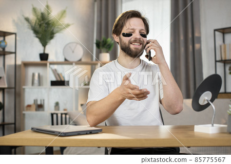 Caucasian bearded man with black collagen hydrogel eye patches talking on cellphone Caucasian bearded man with black collagen hydrogel eye patches talking on cellphone 85775567