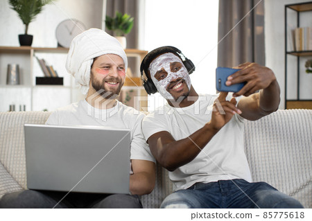 Multiethnic men making facial hygiene procedures, using eye patches and facial mask making selfie 85775618