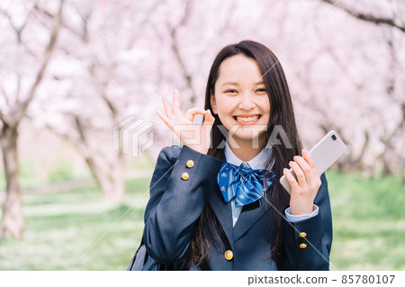 A high school student holding a smartphone under a cherry tree and signing an okay sign 85780107