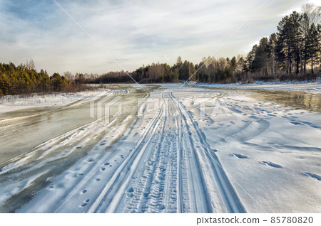 Winter landscape on a sunny day. Ski and snowmobile road along the river. Human footprints. The ice on the river has melted. Trees and bushes grow along the banks of the river. Beautiful sky  85780820
